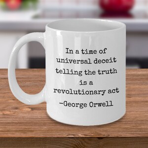 Puede incluir: Taza de café de cerámica blanca con una cita en negro que dice "En tiempos de engaño universal, decir la verdad es un acto revolucionario -George Orwell".