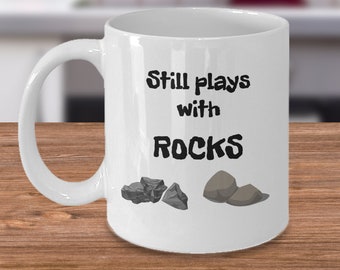 May include: White ceramic coffee mug with black text that reads "Still plays with ROCKS" and a graphic of three grey rocks.