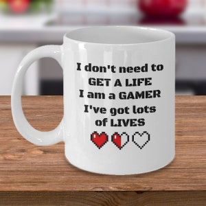 May include: White ceramic coffee mug with black text that reads "I don't need to GET A LIFE I am a GAMER I've got lots of LIVES" with three pixelated red hearts at the bottom.