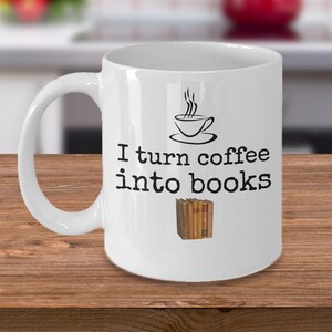 May include: White ceramic coffee mug with black text that reads "I turn coffee into books". A small black and white illustration of a coffee cup with steam rising from it is above the text. A stack of books is to the right of the text.