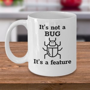 Puede incluir: Taza de cerámica blanca con un gráfico negro de un insecto y el texto "It's not a BUG, It's a feature". La taza tiene un asa curva y está sobre una superficie de madera.