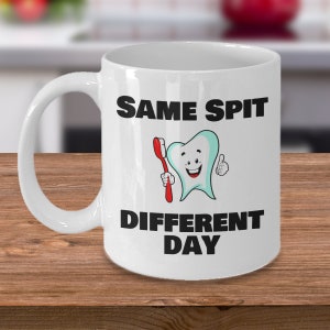 May include: White ceramic coffee mug with a black graphic of a smiling tooth holding a toothbrush. The text "SAME SPIT DIFFERENT DAY" is printed in black.