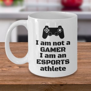 Könnte beinhalten: Weiße Keramik-Kaffeetasse mit einem schwarzen Videospiel-Controller-Motiv und dem Text "Ich bin kein GAMER Ich bin ein ESPORTS-Athlet".