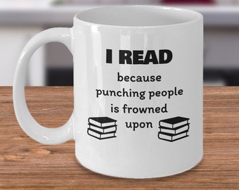 May include: White ceramic mug with the text "I READ because punching people is frowned upon" in black. Two stacks of books are printed on the mug. The mug has a curved handle and is on a wooden surface.