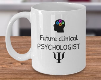 May include: White ceramic mug featuring a black silhouette of a head with colourful gears inside. The text "Future clinical PSYCHOLOGIST" and a black Greek letter psi symbol are printed on the mug.