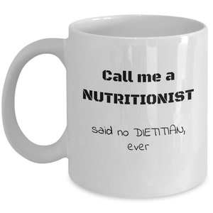 May include: White ceramic coffee mug with black text that reads "Call me a NUTRITIONIST said no DIETITIAN, ever".