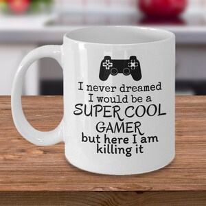 May include: White ceramic coffee mug with a black gamer controller graphic and the text "I never dreamed I would be a SUPER COOL GAMER but here I am killing it".