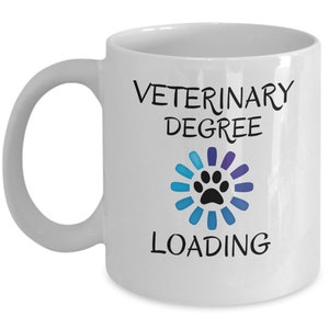 Puede incluir: Taza de café de cerámica blanca con texto negro que dice "Veterinary Degree Loading" con un círculo de carga azul y morado con una huella de pata negra en el centro.