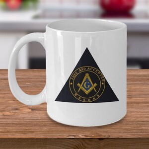 May include: White ceramic mug with a black triangle design featuring a gold and blue Masonic symbol. The symbol includes a compass and square with a letter G in the center. The text around the symbol reads "Free and Accepted Masons".