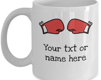 Puede incluir: Taza de café de cerámica blanca con un gráfico de guante de boxeo rojo a cada lado. El texto "Your txt or name here" está impreso debajo de los guantes.