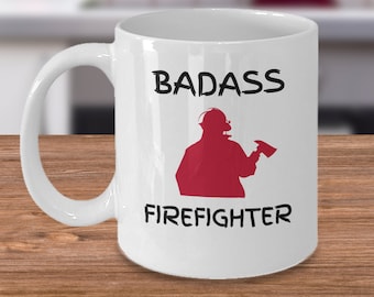 Puede incluir: Taza de café de cerámica blanca con una silueta roja de un bombero sosteniendo un hacha. El texto "BADASS FIREFIGHTER" está impreso en negro en la taza.