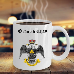 May include: White ceramic coffee mug with a black and gold design. The mug features a double-headed eagle with a crown and the text "Ordo ab Chao".