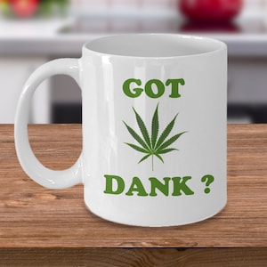 Può includere: Tazza in ceramica bianca con una foglia di cannabis verde e la scritta "GOT DANK ?". La tazza ha un manico curvo. Lo sfondo è una cucina sfocata.