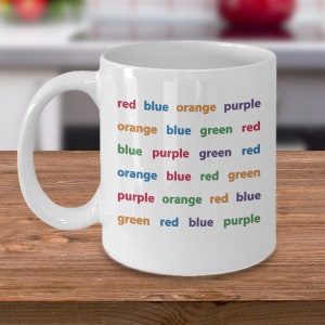 Può includere: Tazza in ceramica bianca con le parole "red blue orange purple" e altre combinazioni di colori stampate in un arcobaleno di colori. La tazza è su una superficie di legno.