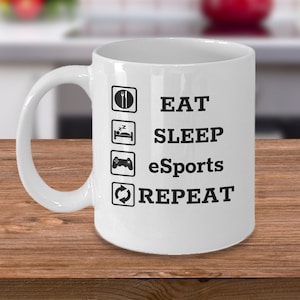 Peut inclure: Une tasse à café en céramique blanche avec un motif noir qui dit "EAT SLEEP eSports REPEAT" avec des icônes pour chaque mot.