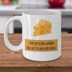 Puede incluir: Taza de cerámica blanca con una cuña de queso amarilla y el texto "Todavía no he encontrado un problema que no se pueda resolver con queso."