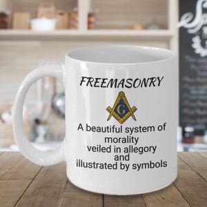 Op de afbeelding: Witte keramische mok met een zwart en gouden vrijmetselaarsymbool en de tekst "FREEMASONRY A beautiful system of morality veiled in allegory and illustrated by symbols".