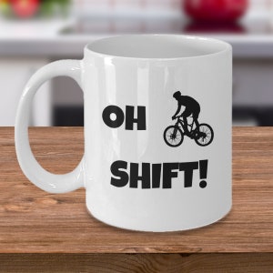 Könnte beinhalten: Weiße Keramik-Kaffeetasse mit einer schwarzen Silhouette eines Radfahrers auf einem Fahrrad und dem aufgedruckten Text "OH SHIFT!"