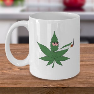 以下が含まれることがあります： 緑色のマリファナの葉のデザインが施された白いセラミック製のマグカップ。葉は赤い目をした漫画の顔をしていて、ジョイントを持っています。