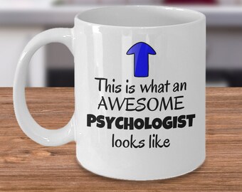 Puede incluir: Taza de café de cerámica blanca con una flecha azul apuntando hacia arriba y el texto "This is what an AWESOME PSYCHOLOGIST looks like".