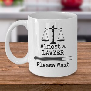 May include: White ceramic coffee mug with a black and white graphic of a scale of justice and the text "Almost a LAWYER Please Wait" with a progress bar underneath.