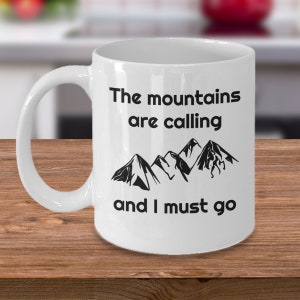 Pode incluir: Caneca de cerâmica branca com um gráfico preto de montanhas e o texto "The mountains are calling and I must go."