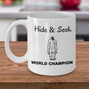 May include: White ceramic coffee mug with a black illustration of a Bigfoot and the text "Hide & Seek World Champion".