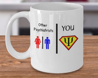 Puede incluir: Taza de café de cerámica blanca con un gráfico en blanco y negro. El gráfico muestra dos figuras de palo que representan a otros psiquiatras y un símbolo de superhéroe que representa "YOU". El texto dice "Other Psychiatrists" y "YOU".
