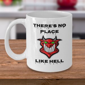 Puede incluir: Taza de café de cerámica blanca con una cara de diablo roja y el texto "There's no place like hell" en letras negras que gotean.