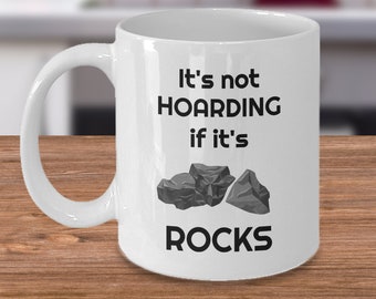 May include: White ceramic coffee mug with a black graphic that reads "It's not hoarding if it's rocks" and a cartoon image of three grey rocks.