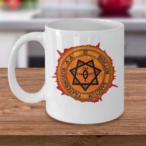 May include: White ceramic mug with a black and orange design. The design features a circle with the text "FRATERNITATIS A.A... SIGILLVM SANCTVM" and a star with the text "7+7 77+77 7" inside.