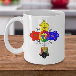 May include: White ceramic mug with a colorful cross design featuring a circle with numbers and symbols. The cross is surrounded by a starburst pattern.