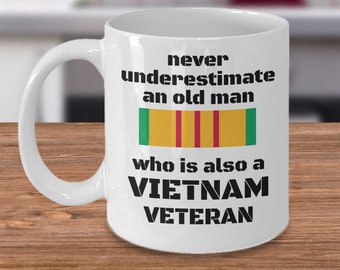 May include: White ceramic coffee mug with a black graphic that reads "never underestimate an old man who is also a VIETNAM VETERAN". The graphic also includes a yellow, red, and green ribbon.