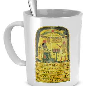 May include: White ceramic mug with a colorful image of an ancient Egyptian stele. The stele depicts a seated figure with a headdress and a standing figure with a staff. Hieroglyphs are inscribed on the stele.