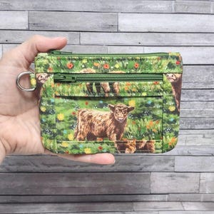 May include: Green fabric coin purse with a zipper closure and a silver-colored ring. The purse features a print of a brown Highland cow in a field of green grass and colorful flowers.
