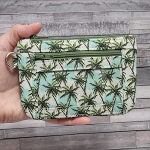 May include: A small, rectangular wallet with a tropical palm tree pattern. The wallet features a light blue background with green palm trees. It has two zippered compartments and a silver key ring attached to the side.