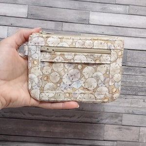 May include: A small, rectangular wallet with a zipper closure and a key ring. The wallet features a beige background with a pattern of sand dollars in shades of beige, tan, and white. The zipper and key ring are silver.