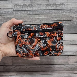 May include: Black wallet with a snake and leaf pattern in orange and white. The wallet has a black zipper and a black ring attached to the side. The wallet is held in a hand.