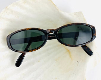 May include: Oval sunglasses with a tortoiseshell pattern frame and dark green lenses. The sunglasses are on a white, textured surface. The text "HAND" is visible on the frame.