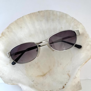 Peut inclure: Lunettes de soleil ovales argentées avec des verres gris foncé. Les lunettes de soleil reposent sur une coquille de coquillage blanche.