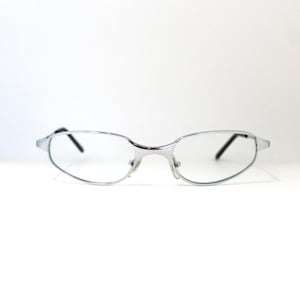May include: Silver-framed eyeglasses with oval lenses. The glasses have a thin, metallic frame and black temples. The lenses are clear, and the overall design is sleek and modern. The glasses are set against a white background.