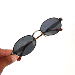 May include: Oval-shaped sunglasses with dark gray lenses and a black metal frame. The temples are a tortoiseshell pattern with a silver bridge. The sunglasses are held against a white background.