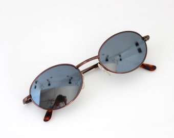 May include: Oval-shaped sunglasses with a brown metal frame and blue-tinted lenses. The temples have a tortoiseshell pattern. The sunglasses are set against a white background.