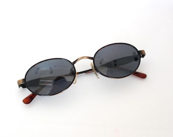 May include: Oval-shaped sunglasses with dark gray lenses and a thin, bronze-colored metal frame. The temples have a reddish-brown hue. The sunglasses are designed in a classic style.