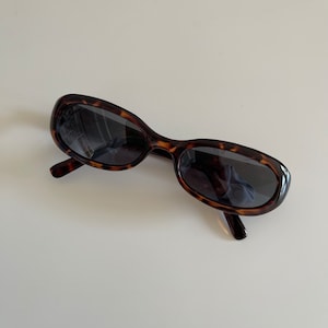 Peut inclure: Lunettes de soleil ovales avec un motif écaille de tortue. Les montures sont marron et noires, et les verres sont foncés. Les lunettes sont posées sur une surface blanche.