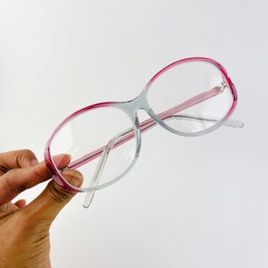 Könnte beinhalten: Klare Plastikbrille mit rosafarbenem Rand. Die Brille hat eine abgerundete, ovale Form.