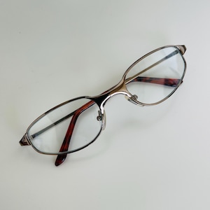 May include: A pair of eyeglasses with a gold-colored metal frame and clear lenses. The temples are a dark reddish-brown color with a tortoiseshell pattern. The glasses are designed with a sleek, modern aesthetic.
