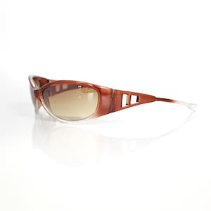May include: Brown-framed sunglasses with a gradient lens. The sunglasses have a rectangular shape with a clear lower frame and a brown upper frame. The arms of the sunglasses have a cut-out design.
