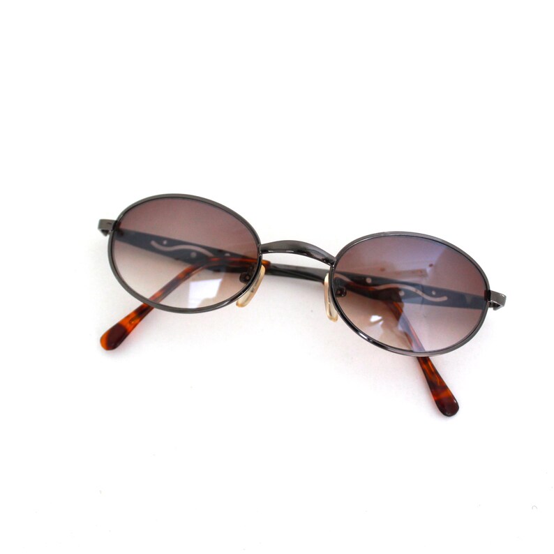 Pode incluir: Um par de &oacute;culos de sol ovais com lentes de degrad&ecirc; marrom e arma&ccedil;&atilde;o de metal preto. Os &oacute;culos t&ecirc;m um padr&atilde;o de tartaruga nos bra&ccedil;os.