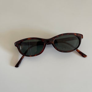 May include: Tortoise shell sunglasses with dark lenses. The sunglasses have a rounded, oval shape.